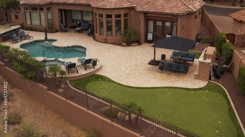 An aerial 4k video of a Deseret landscaped backyard in Arizona featuring a travertine tiled pool deck with a spa, outdoor fireplace, outdoor kitchen and putting green.