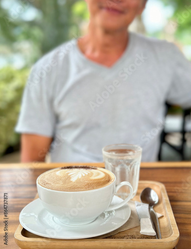 A fresh cappuccino coffee in white round porcelain cup and plate with stainless steel spoon and sugar pack and pure water in the natural wood tray on wood with a man smiling in background
