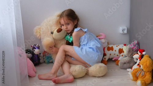A charming little girl with pigtails among soft toys at home is sitting on the floor sad, hugging a big bear. 