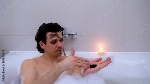 Man applying charcoal face mask lying in bath with foam and candle at home. Spa, hygiene, skin care procedure, treatment, wellness. Handsome young brunette male relaxing in bathroom in evening.