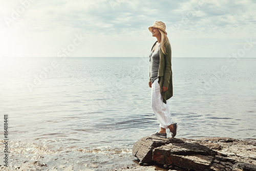 Happy woman, thinking and beach with sunshine in nature for outdoor holiday, weekend or journey. Young female person on rock with smile for travel, vacattion or freedom by ocean coast, water or sea photo