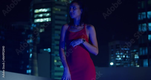 A young girl in a red dress is on a rooftop at night, city buildings looming in the background. photo