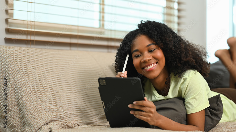 Happy young African American woman lying on couch at home using digital tablet Happy young African American woman lying on couch at home using digital tablet