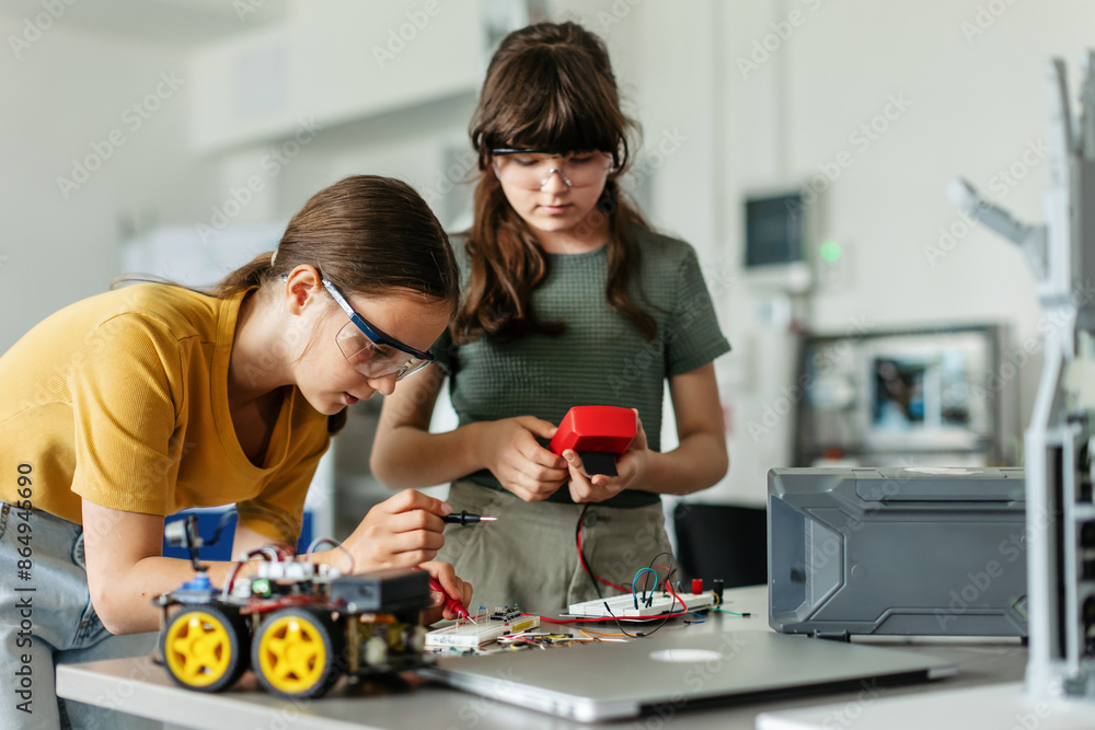 Fototapeta premium Two girl classmates working togeter on circuit board, building robotic car in after-school robotics club. Children learning robotics in Elementary school.