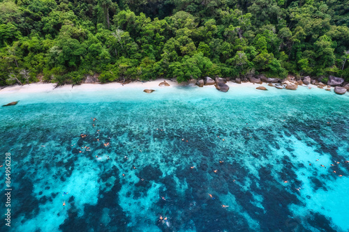 Aerial view of a pristine tropical beach with turquoise waters and lush green forest in the background, swimmers enjoying the clear blue sea © escapejaja