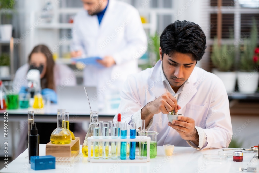 Focus at Young Scienctist man using grinding bow with experiment tube with blue liquid with intense expression face. With blur his co-workers working behide. Focus at Young Scienctist man using grinding bow with experiment tube with blue liquid with intense expression face. With blur his co-workers working behide.