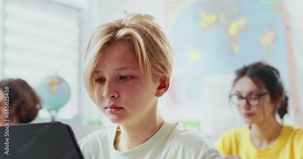 Camera focus on Caucasian boy. Pupil sitting with classmates during lesson. Watching video or completing test on tablet device. Modern technology concept. Attentive child learning in school. Camera focus on Caucasian boy. Pupil sitting with classmates during lesson. Watching video or completing test on tablet device. Modern technology concept. Attentive child learning in school.
