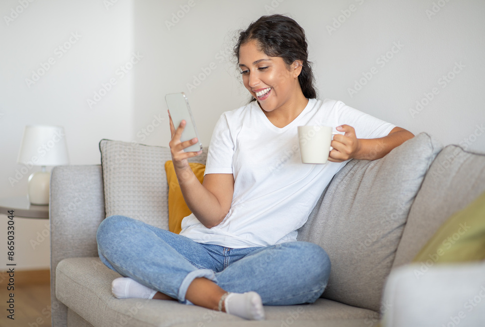 A woman sits on a light-colored sofa in a living room. She wears a white t-shirt and blue jeans and smiles while looking at her phone with a cup of coffee in her hand. A woman sits on a light-colored sofa in a living room. She wears a white t-shirt and blue jeans and smiles while looking at her phone with a cup of coffee in her hand.