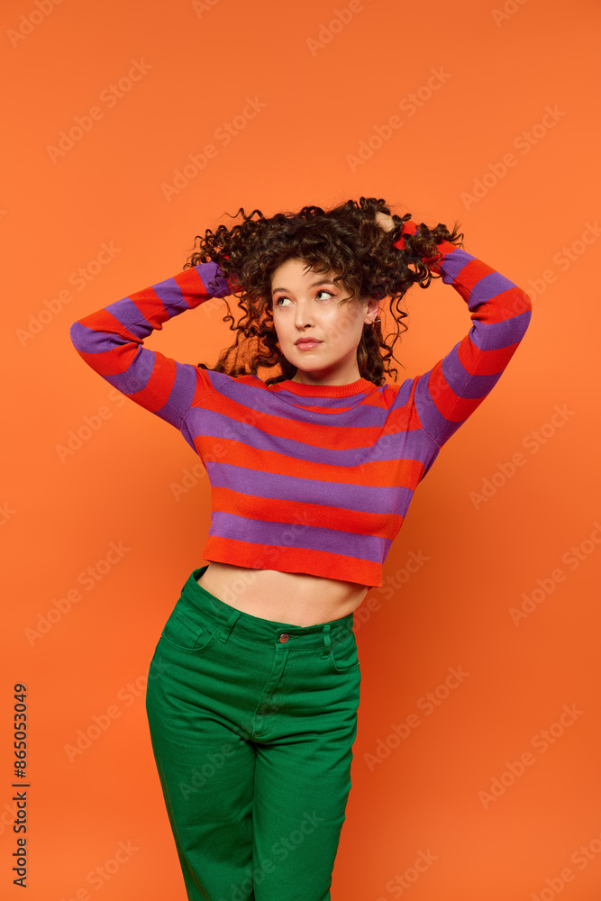 A young woman with curly brown hair poses against a bright orange backdrop wearing a red and purple striped sweater and green pants. A young woman with curly brown hair poses against a bright orange backdrop wearing a red and purple striped sweater and green pants.