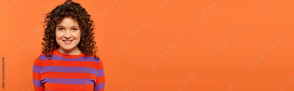 A young woman with curly hair smiles brightly while posing in vibrant clothing against a bold orange background. A young woman with curly hair smiles brightly while posing in vibrant clothing against a bold orange background.