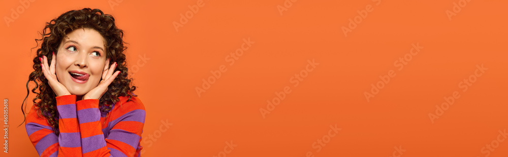 A young woman with curly hair smiles and sticks her tongue out while posing against a bright orange background. A young woman with curly hair smiles and sticks her tongue out while posing against a bright orange background.