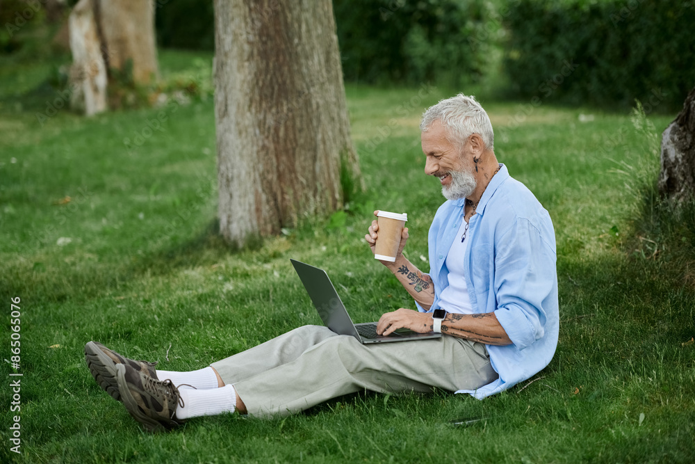 A mature man with tattoos and a grey beard sits on the grass in a park, working on his laptop while holding a coffee cup. A mature man with tattoos and a grey beard sits on the grass in a park, working on his laptop while holding a coffee cup.