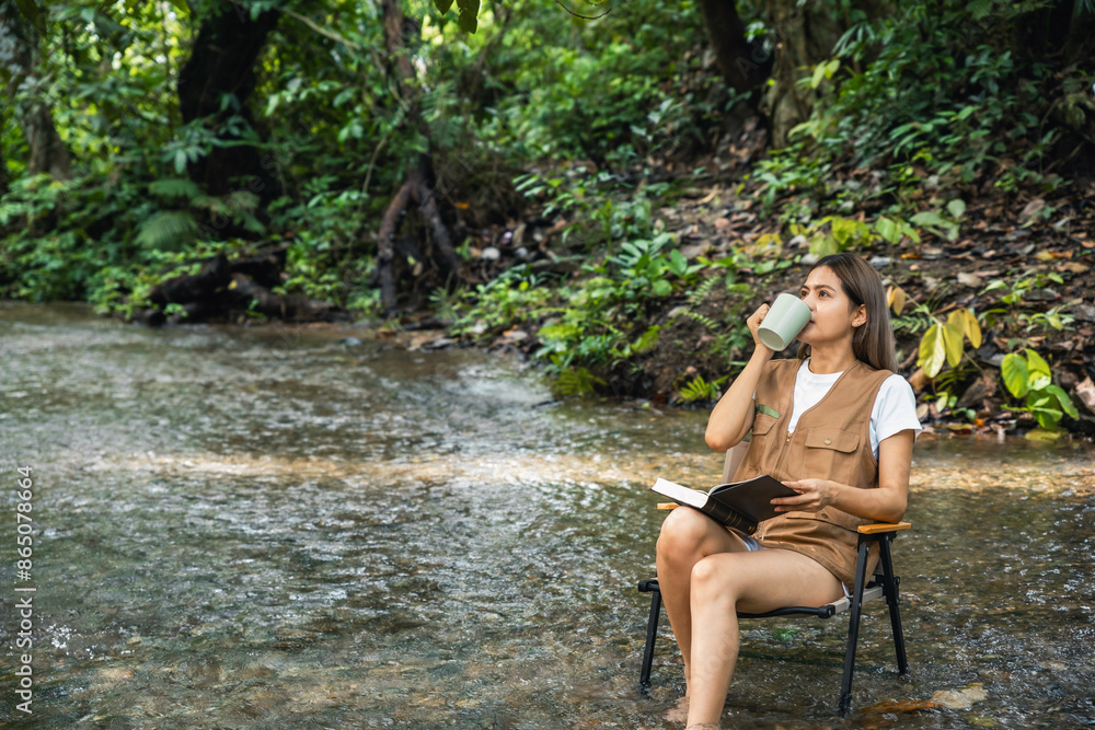 Fototapeta premium Beautiful happy young Asian woman sitting on camping chair drink coffee and reading a book in middle of a river in park on vacation. copy space on the left.