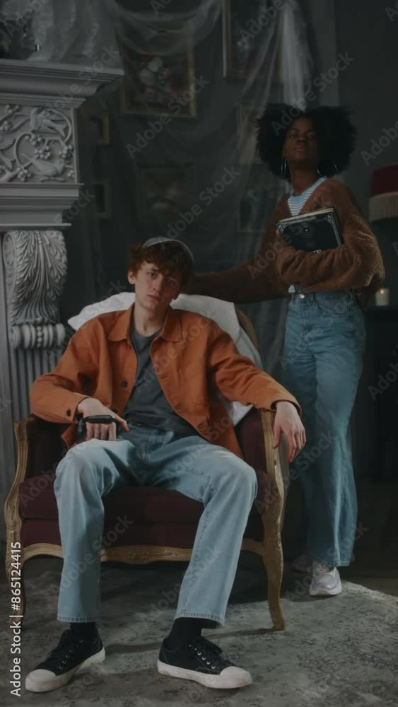 Vertical full shot of cool Caucasian guy in casual clothes and cap posing in armchair and African American lady by him with occult book in old desolate mansion Vertical full shot of cool Caucasian guy in casual clothes and cap posing in armchair and African American lady by him with occult book in old desolate mansion