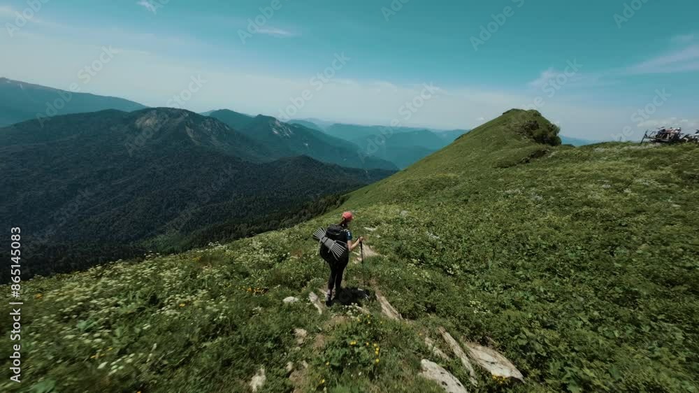 A girl with a hiking backpack and trekking poles walks along the top of a picturesque mountain in green grass surrounded by clouds and high mountains covered with snow A girl with a hiking backpack and trekking poles walks along the top of a picturesque mountain in green grass surrounded by clouds and high mountains covered with snow