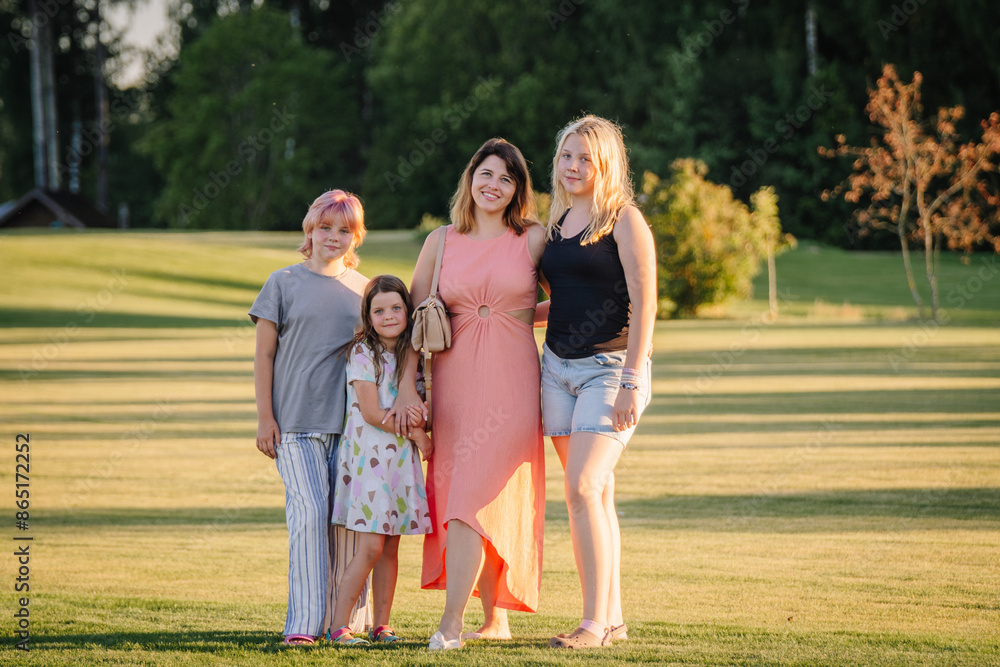 Obraz premium Valmiera, Riga - June 26, 2024 - A woman with three children posing in a grassy field, smiling and looking at the camera, with trees and a building in the background, ample copy space.