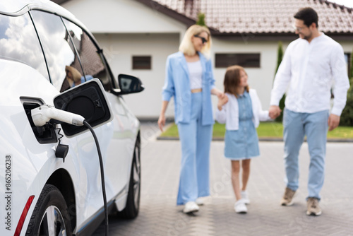 A Family Stands Near Electric Car Being Charged Outside Home, Emphasizing Eco-Friendly Living And Modern Technology
