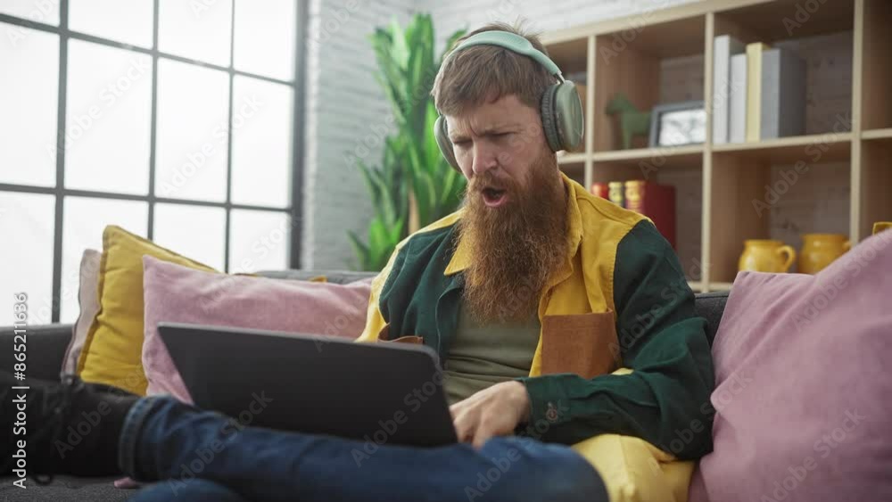 A cheerful bearded redhead man in casual clothes enjoys music on headphones while using a laptop on a comfy sofa indoors. A cheerful bearded redhead man in casual clothes enjoys music on headphones while using a laptop on a comfy sofa indoors.