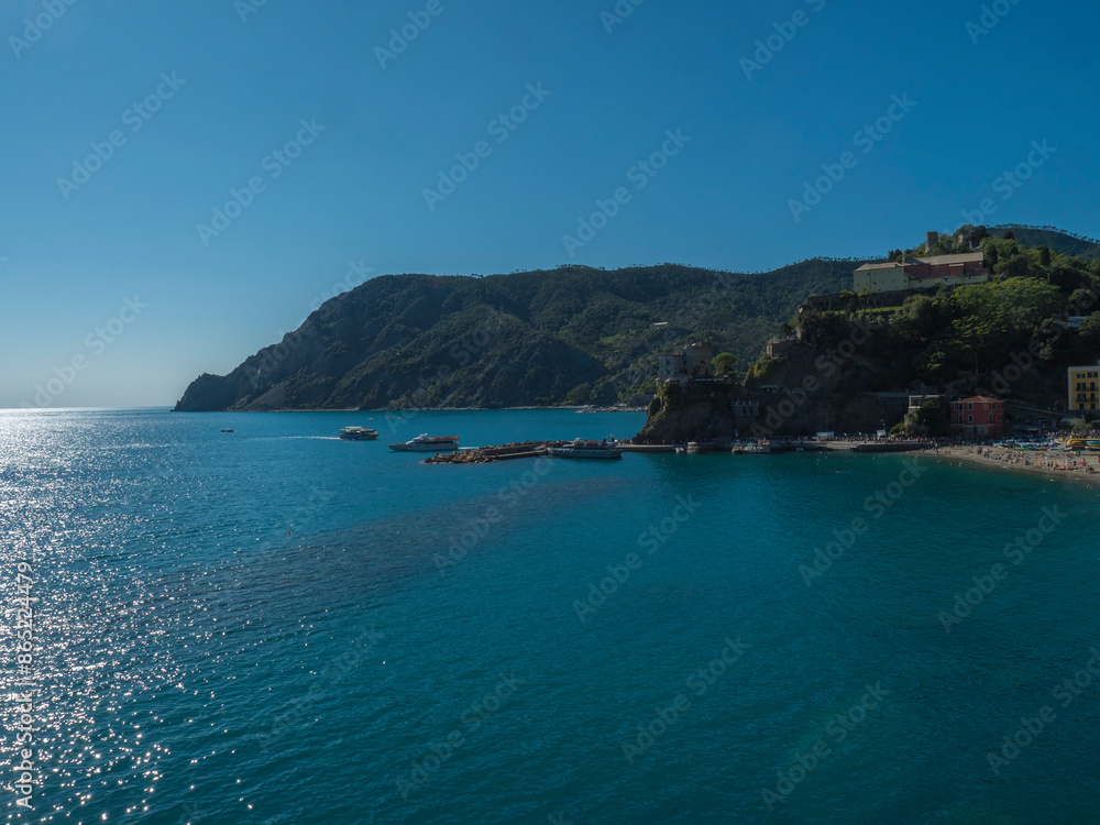 Fototapeta premium Picturesque distant view of Monterosso al Mare with beach, ships and boats seen from Azure Trail to Vernazza with green cliffs, rock, clear blue sky and sea. National park Cinque Terre, Liguria, Italy