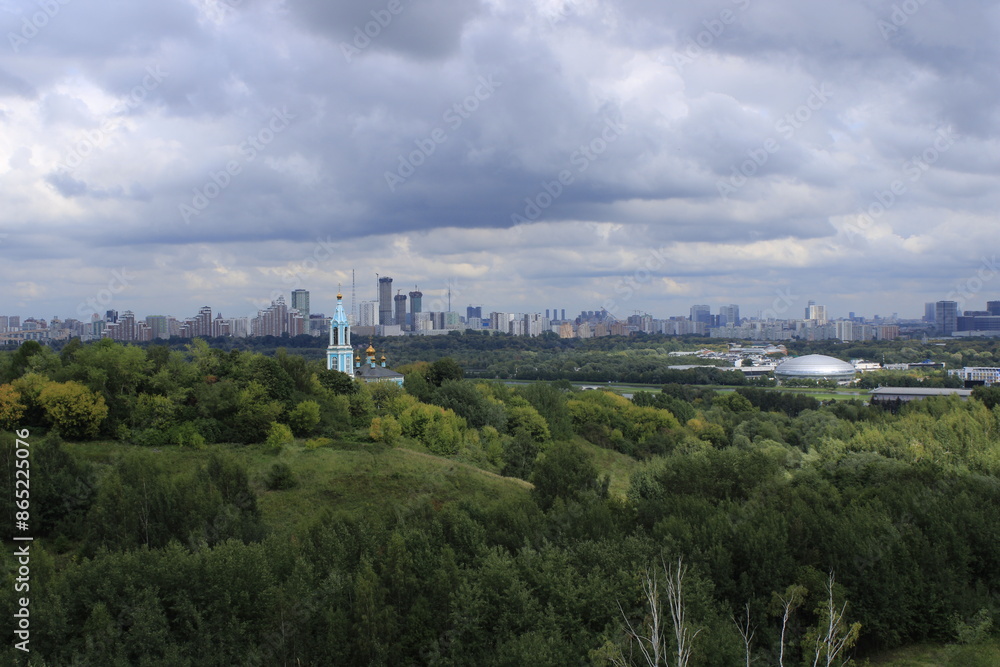 A serene landscape captures the harmony between nature and urban life. The vibrant green trees and a charming blue church contrast with the modern city skyline under a moody, cloudy sky A serene landscape captures the harmony between nature and urban life. The vibrant green trees and a charming blue church contrast with the modern city skyline under a moody, cloudy sky
