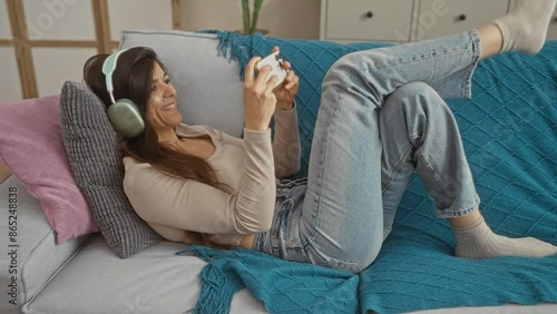 Young woman playing video games on her smartphone while wearing headphones and relaxing on a couch in a cozy living room. photo