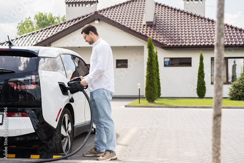 Man Charging Electric Car At Home In Driveway On A Sunny Day. Sustainable Technology, Eco-Friendly Transportation, Green Energy Concept.