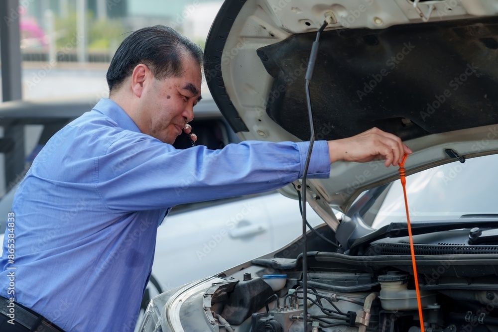 Asian insurance officer inspecting car engine while talking on the phone. Wearing blue shirt. Represents insurance claim processing, customer service, and vehicle inspection in the automotive industry Asian insurance officer inspecting car engine while talking on the phone. Wearing blue shirt. Represents insurance claim processing, customer service, and vehicle inspection in the automotive industry