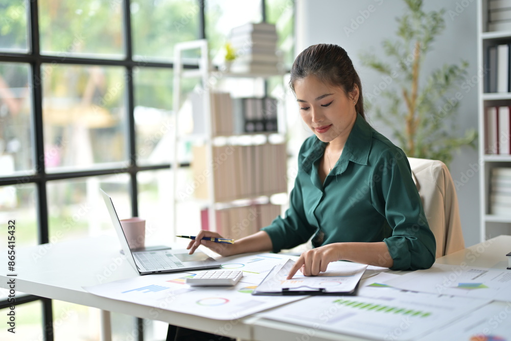 Focused young woman working at office desk with laptop, analyzing financial charts, showcasing dedication in modern corporate setting Focused young woman working at office desk with laptop, analyzing financial charts, showcasing dedication in modern corporate setting