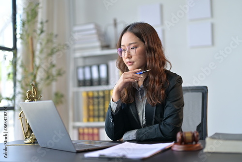 Asian lawyer focused on her laptop in an office, surrounded by law symbols, reviewing legal documents. The scene exudes business and justice