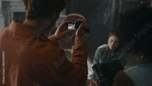 Back view of guy filming video on vhs camera of writhing demon possessed girl tied up to chair and of lady with occult book searching for prayers to perform exorcism ceremony in old derelict house