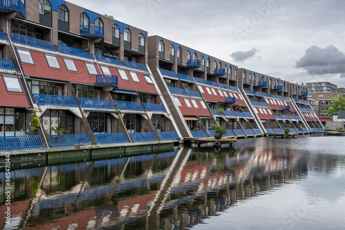 A row of modern, multi-story buildings with vibrant red and blue accents reflecting in a still canal, showcasing contemporary architecture alongside serene water imagery in Rotterdam photo