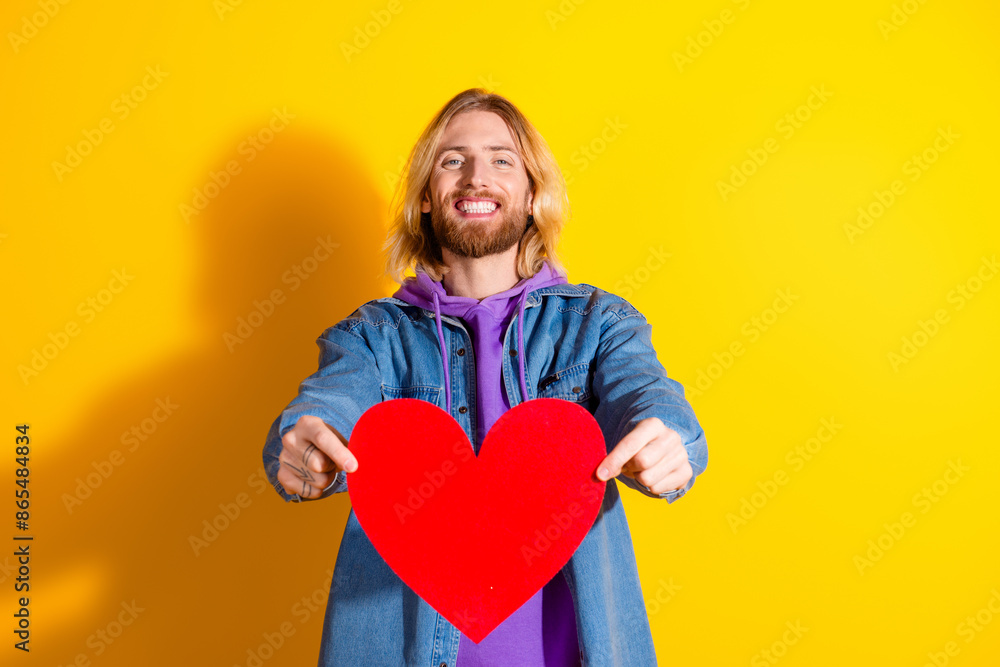 Photo portrait of attractive young man hold big red heart dressed stylish denim clothes isolated on yellow color background Photo portrait of attractive young man hold big red heart dressed stylish denim clothes isolated on yellow color background
