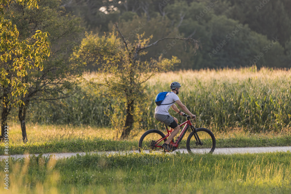 Naklejka premium Man biking electric mountain bike along the trail in a fantastic rural landscape at sunset