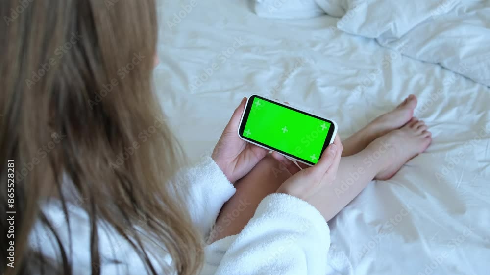The blonde is sitting on the bed. Women's hands hold a phone with a green screen. 4k video. High quality 4k footage The blonde is sitting on the bed. Women's hands hold a phone with a green screen. 4k video. High quality 4k footage