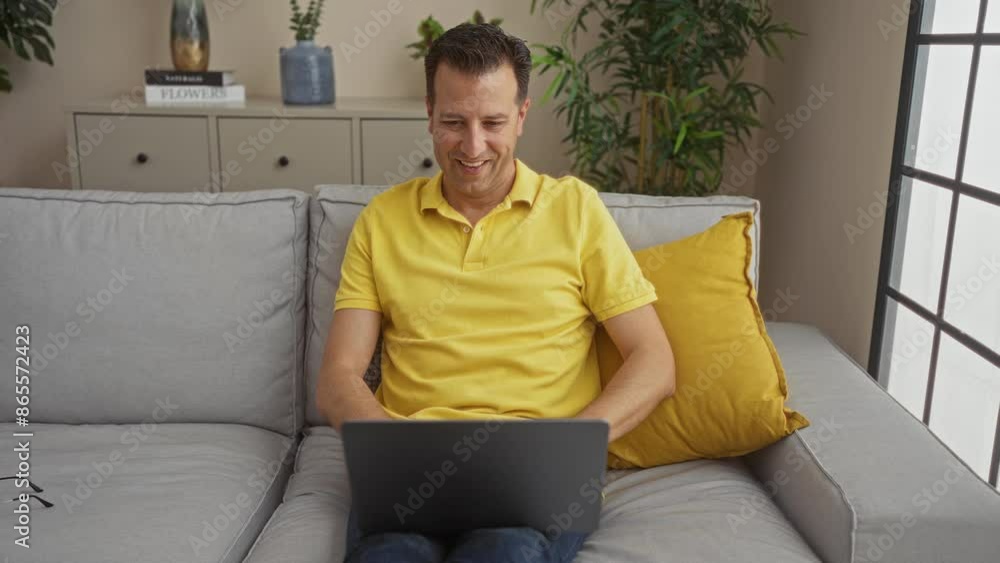 Man working inside home living room using laptop sitting on sofa with plants and decor behind him smiling in a yellow shirt Man working inside home living room using laptop sitting on sofa with plants and decor behind him smiling in a yellow shirt