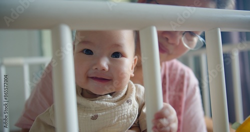 Adorable Baby infant Smiling Through playpen fence with grandmother in Background, Capturing showcasing a tender family Heartwarming Moment of Joy and Innocence in a Cozy Home Environment