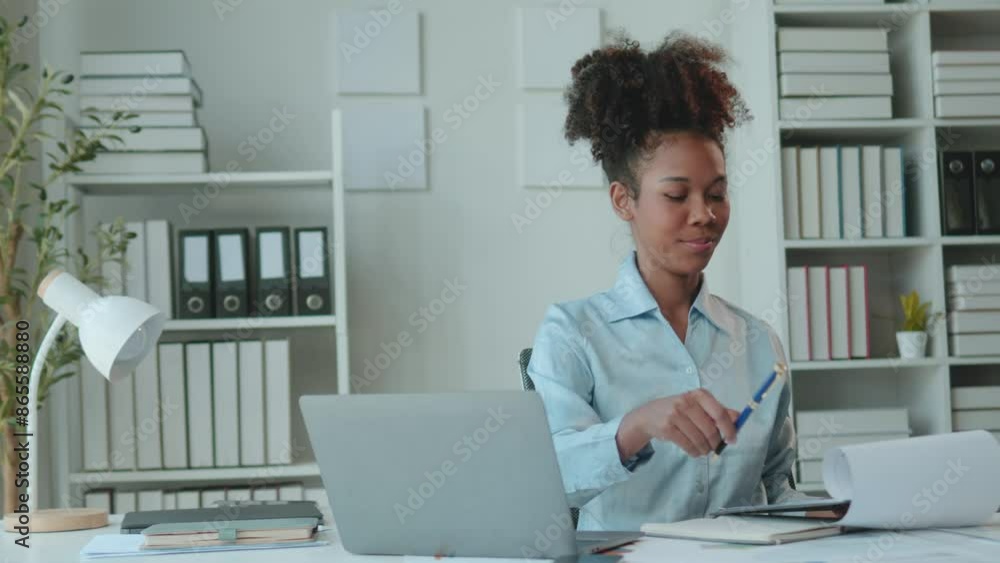 Young african american businesswoman works on a laptop in a modern office, reviewing documents. Shelves and files are in the background Young african american businesswoman works on a laptop in a modern office, reviewing documents. Shelves and files are in the background