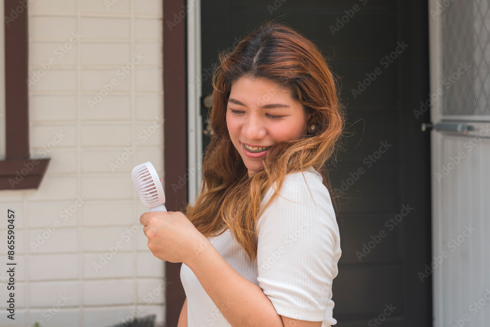 An Asian woman in her 30s cooling off with a handheld portable USB fan outdoors against a house background. An Asian woman in her 30s cooling off with a handheld portable USB fan outdoors against a house background.