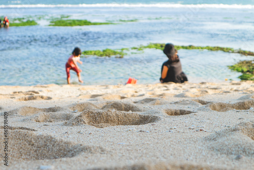 Seascape abstract beach background, child and their mother playing in the water. Blurred bokeh light on the sea. Focus on the sand foreground.