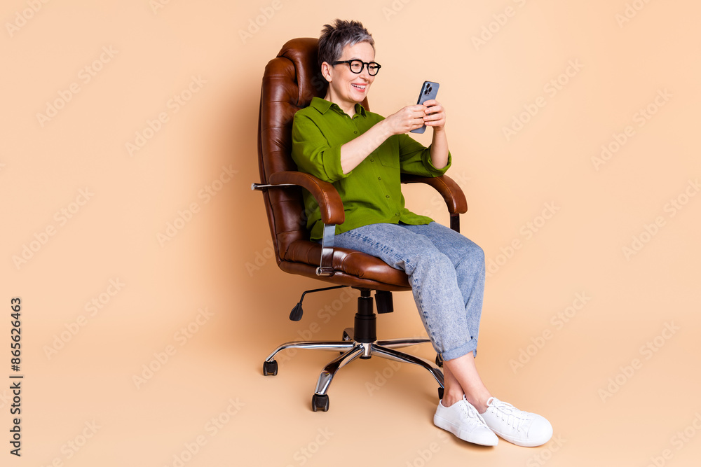 Full body photo of attractive retired woman sit armchair hold device dressed stylish green clothes isolated on beige color background Full body photo of attractive retired woman sit armchair hold device dressed stylish green clothes isolated on beige color background