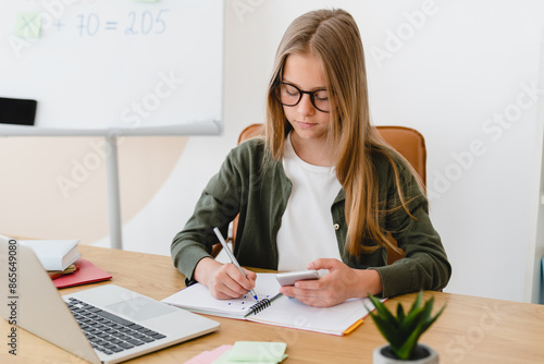 Fotografía Smart caucasian student teenager schoolgirl pupil e-learning, doing homework projects remotely, preparing for test, tutoring online from home using laptop and smart phone indoors