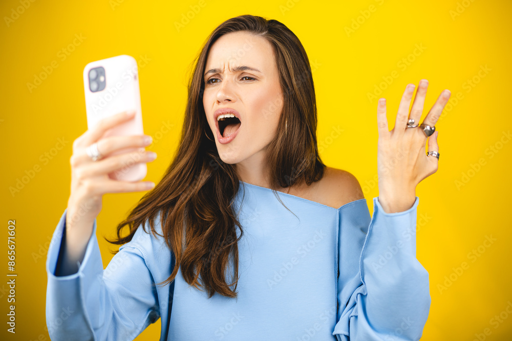 Mad and shock young brunette beautiful woman using mobile phone isolated on yellow background. Yelling unhappy woman hold mobile phone. Disappointed sad upset lady horrified impressed news. WTF. Oh no Mad and shock young brunette beautiful woman using mobile phone isolated on yellow background. Yelling unhappy woman hold mobile phone. Disappointed sad upset lady horrified impressed news. WTF. Oh no