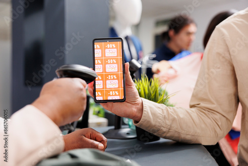 Digital coupon marketing. Customer using mobile discount code while shopping in clothing store on Black Friday, close up of man showing smartphone with digital voucher to retail employee at checkout photo