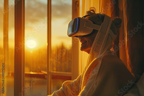 A woman is sitting in front of a window, wearing a VR headset. The sun is setting in the background, creating a warm and cozy atmosphere. The woman is enjoying her virtual reality experience