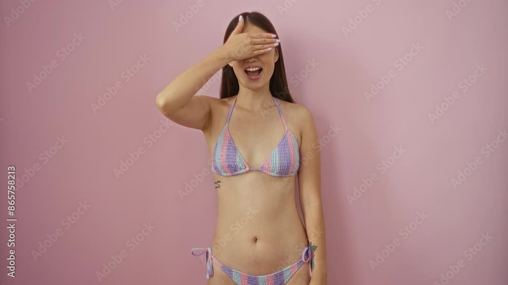 Smiling young chinese woman in bikini covering eyes with hand for a surprising blind expression over isolated pink background Smiling young chinese woman in bikini covering eyes with hand for a surprising blind expression over isolated pink background