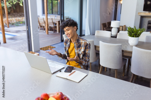 Video calling on laptop, teenage boy gesturing and smiling in modern kitchen, copy space