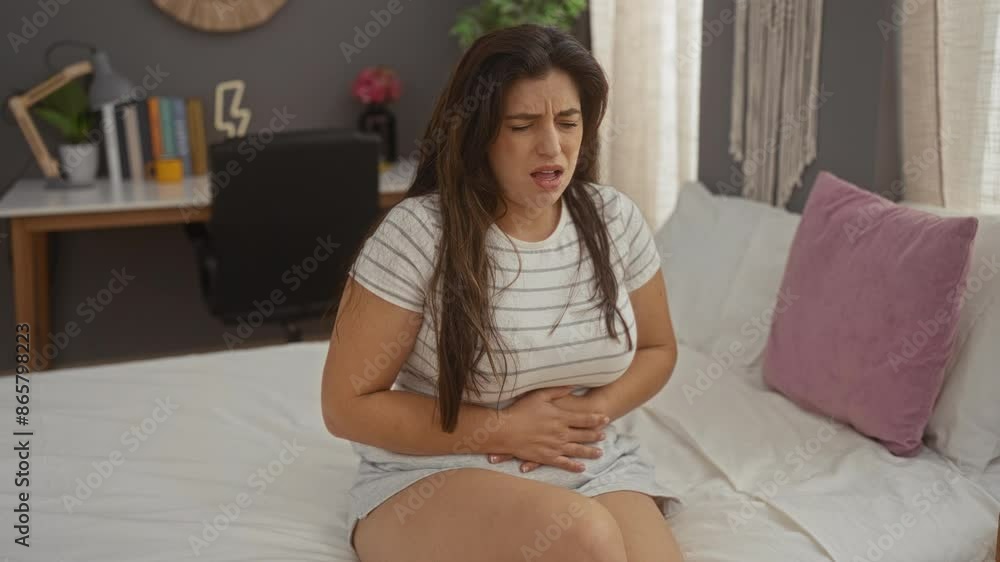 Woman in casual attire experiencing stomach pain while sitting on bed in a cozy bedroom setting with desk and plants in the background Woman in casual attire experiencing stomach pain while sitting on bed in a cozy bedroom setting with desk and plants in the background