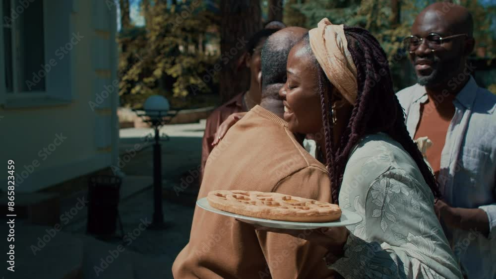 Medium close-up slowmo shot of cheerful young African American woman with braids, holding cake hugging senior father while smiling family is watching, during visit to celebrate holiday Medium close-up slowmo shot of cheerful young African American woman with braids, holding cake hugging senior father while smiling family is watching, during visit to celebrate holiday