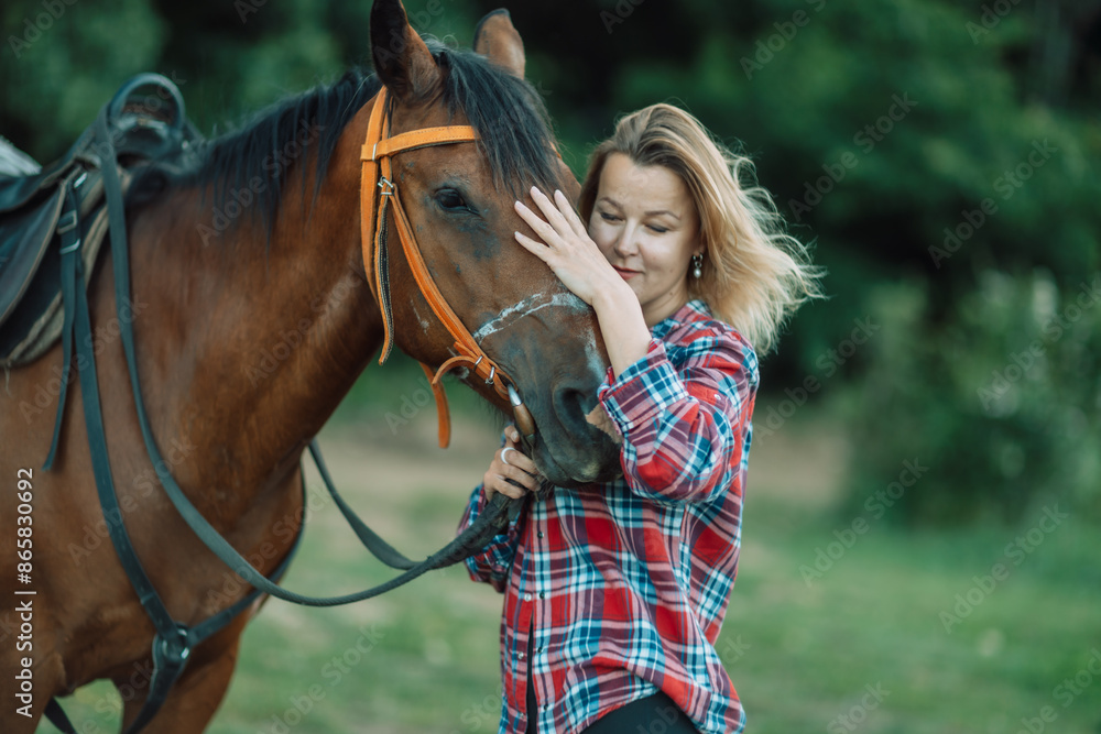 Obraz premium Happy blonde with horse in forest. Woman and a horse walking through the field during the day. Dressed in a plaid shirt and black leggings.
