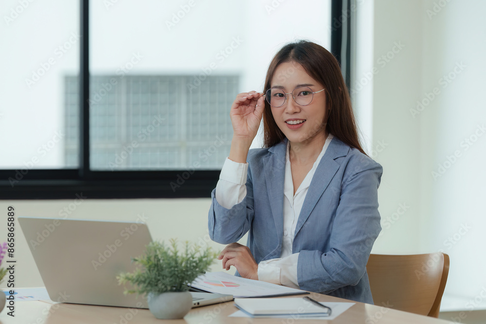 A woman in a business suit sits at a desk with a laptop and a stack of papers A woman in a business suit sits at a desk with a laptop and a stack of papers
